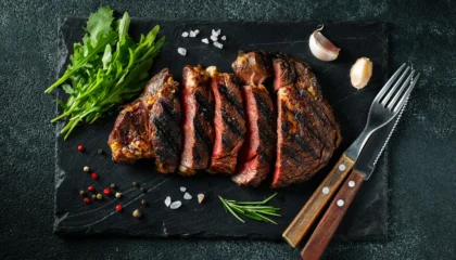 Sliced steak ribeye, grilled with pepper, garlic, salt and thyme served on a slate chopping Board on a dark stone background. Top view. Flat lay.